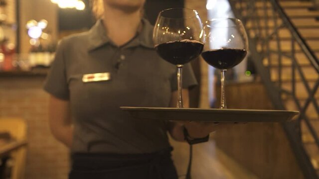 Young waitress in grey polo shirt carries tray with glasses of delicious red wine to guests sitting at table in comfortable restaurant close view slow motion.