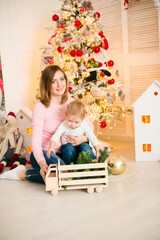 Cute little boy with blond hair plays with his mother in a bright room decorated with Christmas garlands near the Christmas tree. Happy childhood. Christmas mood