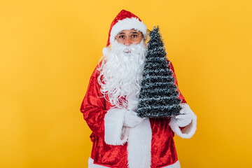 Fototapeta na wymiar Santa Claus holding a Christmas tree on yellow background. Christmas concept
