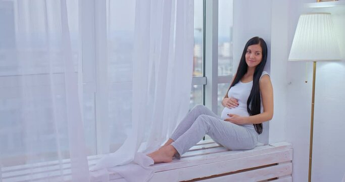 Beautiful pregnant woman is sitting by the window, waiting for the baby. Pregnant woman stroking her tummy with her hand, happy motherhood. 4k, ProRes