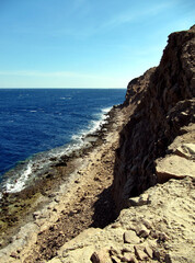 View along the cliff into the abyss of the blue sea with white waves and blue sky.