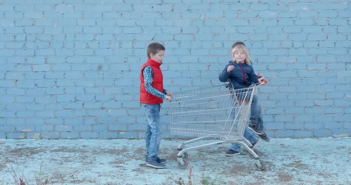 Three funny teenage boys in bright clothes ride little brother on grocery cart in backyard against blue brick wall