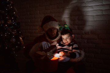A cute smiling boy is sitting in the arms of Santa Claus in the dark and looking at a lighted gift for Christmas Eve late at night. Happy New Year and merry xmas