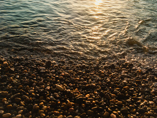 Seashore with pebbles at sunset, the sun reflected in the water.