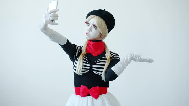 Female mime artist is taking selfie with smart phone camera posing with funny faces then swiping touching screen enjoying photos on white background