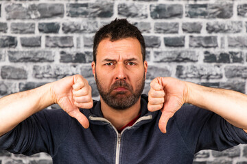 Young caucasian man holding both thumbs down with a frowning face in front of a stone wall