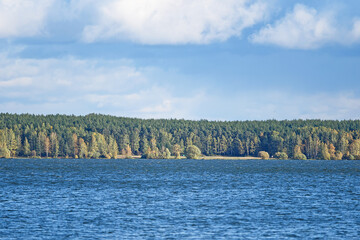 Autumn view from the surface of the lake on the shore overgrown with deciduous forests against a blue sky with white clouds