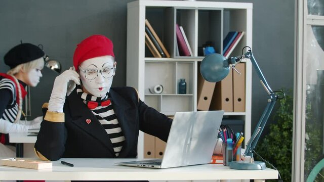 Mimes office workers in funny costume and make-up are talking on mobile phone and using laptops in workplace. Business activities and communication concept.