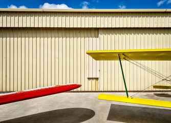 a red wing and a yellow biplane wings against a faded yellow corrugated hanger door and a blue sky with clouds