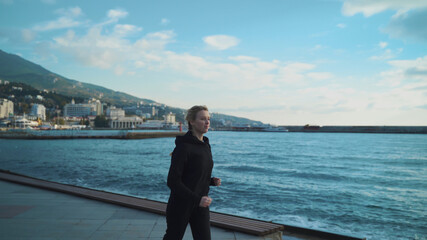 A young blonde girl in the early morning is engaged in jogging along the sea against the background of the lighthouse.