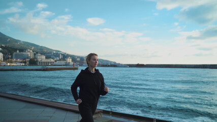 A young blonde girl in the early morning is engaged in jogging along the sea against the background of the lighthouse.