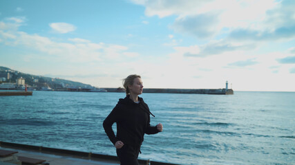 A young blonde girl in the early morning is engaged in jogging along the sea against the background of the lighthouse.