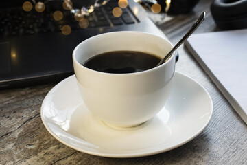 A white Cup of coffee stands on a wooden table against the background of a laptop with bokeh and Notepad. The concept of a break during work and a business meeting