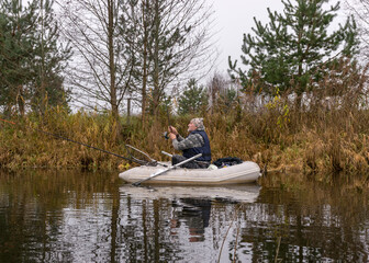 gray and cloudy day, fisherman in a white boat, river bank with bare trees and bushes, shore reflection in river water