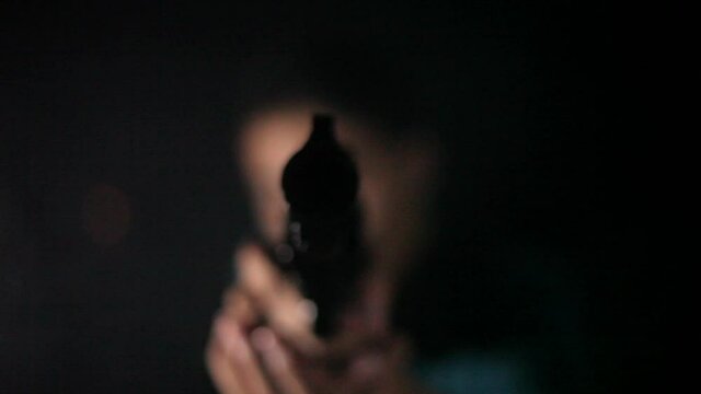 Shot of a man in gree shirt walking and pointing gun towards the camera in dark smoky background.