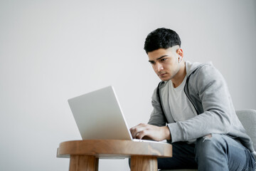 a young man is reading the latest news at home on a laptop computer, doing an Economics assignment on his new project. He is dressed in gray clothing and wears wireless headphones in his ears