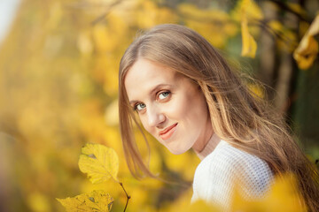 Beautiful young woman with light brown hair in a white sweater on a background of foliage in an autumn park. Portrait of an attractive happy woman among colorful yellow leaves. Fall season. Autumn.