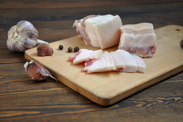 Pieces of pork fat on a wooden board. Garlic and peppercorns. Dark wooden table.