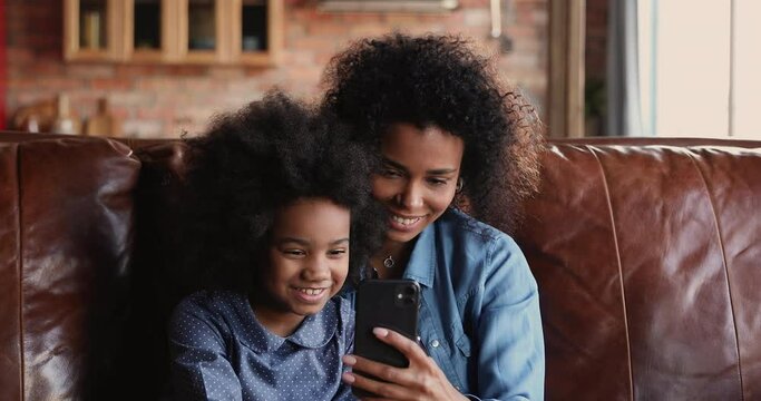 Beautiful african young mom her little daughter using smartphone taking selfie picture, having fun download new funny app, make video call rest on sofa at home. Modern tech, parental control concept