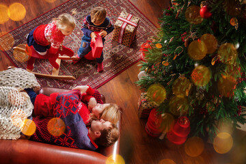 Happy family is spending time together at living room near christmas tree. Parents with children are celebrating new year holidays opening presents. Festive evening at decorated cozy home. Winter mood
