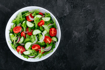Fototapeta na wymiar Healthy green salad with fresh tomato, cucumber, red onion and lettuce in bowl on dark stone background.
