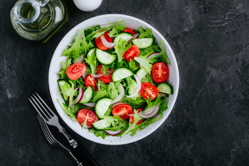 Healthy green salad with fresh tomato, cucumber, red onion and lettuce in bowl on dark stone background.