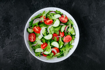Healthy green salad with fresh tomato, cucumber, red onion and lettuce in bowl on dark stone background.