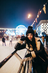 Happy girl in warm clothes stands in the evening near the rink on the background of the Christmas street and people, looks at the camera and smiles. Entertainment on Christmas weekend. Vertical