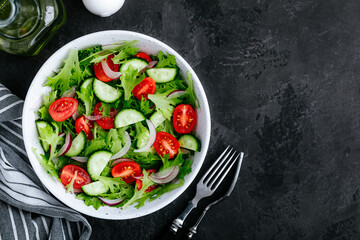 Healthy green salad with fresh tomato, cucumber, red onion and lettuce in bowl on dark stone background.