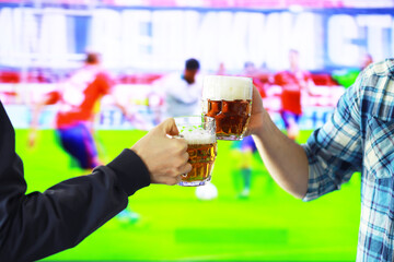 Two hands horizontal holding lager beer glass and clinking on background of football game. Sport fans cheer up. Friends leisure lifestyle concept.