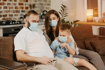 Father, mother and son are sitting on a sofa in medical face masks to avoid the spread of coronavirus (COVID-19). Family during quarantine at home. Relatives are watching video on a cellphone on couch
