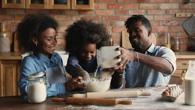 Playful african little 6s daughter prepare dough for pastries with parents at home. Full family gathered in domestic modern cozy kitchen cooking together enjoy weekend making holiday cake or cookies