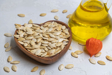 Fried pumpkin seeds lies in heart shaped wooden bowl on the gray background next to glass bottle of...