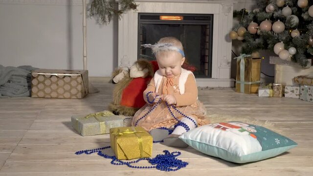 Charming one-year-old girl playing near the Christmas tree with Christmas decorations sitting on the floor. The concept of a happy Christmas. Portrait. 4K