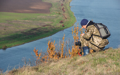 a man photographs the landscape near the river in camouflage clothes with a backpack on his back