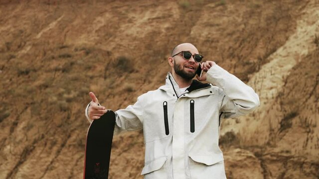 Handsome bald bearded man with sunglasses talks on phone with sandboard in desert or sand quarry