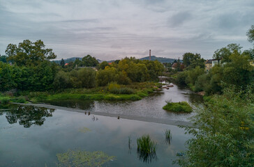 Fototapeta premium Cloudy Landscape of small river between bushes and trees with small waterfall