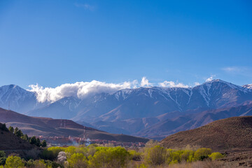 Daytime wide angle shot of Beautiful landscape of snow capped mountains and bushes and a village in the valley. Atlas, Morocco.