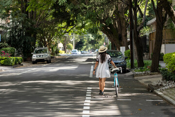 Mujer con vestido y sombrero caminando por la calle llevando una bicicleta, con arboles a los lados en un dia de invierno.
