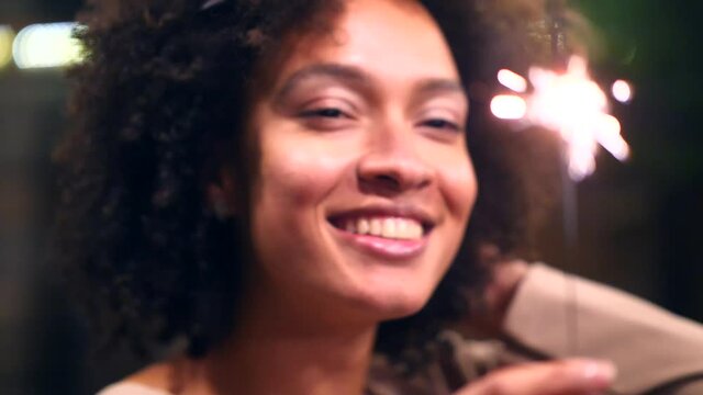 Cheerful young woman holding single sparkler in hand outdoor. Detail of afro american girl celebrating new year’s eve with bengal light. beautiful woman holding a sparkling stick at party night.