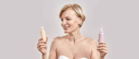 Portrait of beautiful middle aged woman holding two bottles of different cosmetic skincare products and choosing what to apply while posing isolated over grey background