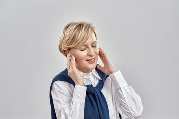 Portrait of elegant middle aged caucasian woman wearing business attire, adjusting her earbuds, enjoying the sound with eyes closed while posing isolated over grey background