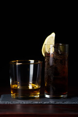 Whiskey neat and cola with ice and lemon wedge served on slate plate. Dark red wooden table, high resolution
