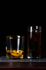 Whiskey with lemon zest and cola with clear ice served on slate plate. Dark red wooden table, high resolution