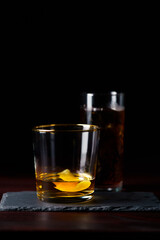 Whiskey with lemon zest and cola with clear ice served on slate plate. Dark red wooden table, high resolution