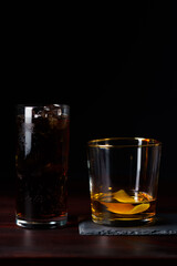 Whiskey with lemon zest and cola with clear ice served on slate plate. Dark red wooden table, high resolution