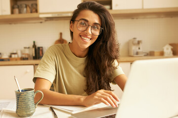 Pretty young woman designer in stylish eyeglasses working from home, sitting at kitchen table with cup of coffee, using high speed internet connection. Cute Hispanic girl shopping online via laptop