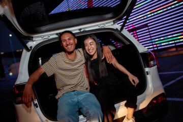 Two young well-dressed friends of different genders and nationalities laughing and sitting together in an opened car trunk outside on a parking site with a led screen behind