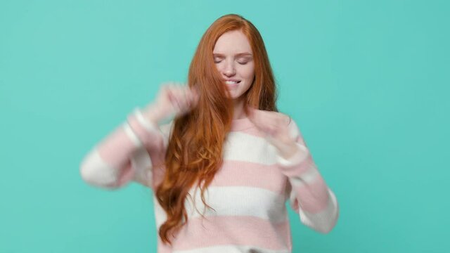 Smiling cheerful pretty young redhead girl in white pink sweater posing isolated on blue turquoise background in studio. People emotions lifestyle concept. Dancing clenching fists rising waving hands