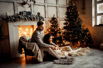 Beautiful couple sit near fireplace and Christmas tree, charming husband hugging adorable wife,...